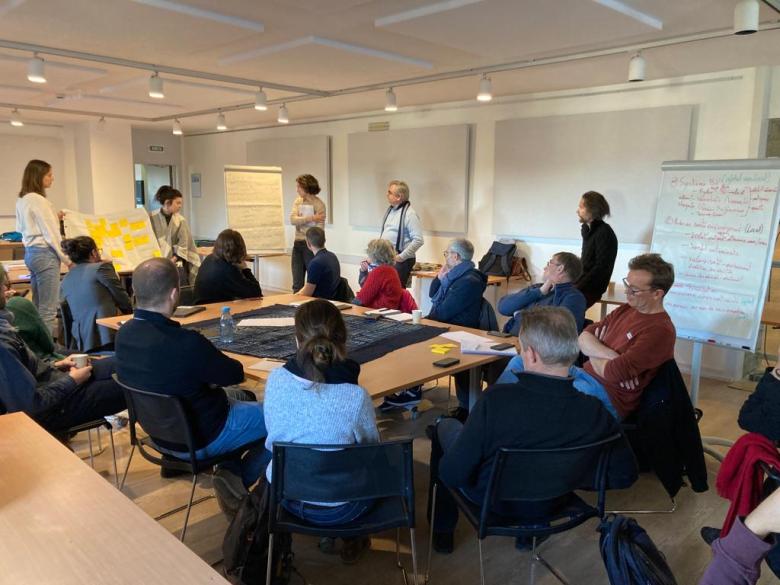 Picture of a group of students and researchers seated around a table listening to the summary of a group workshop